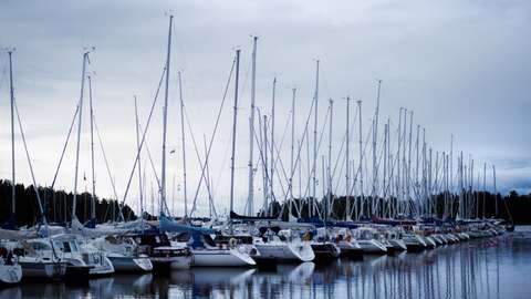 A row of sailboats moored at Finnoonsatama harbour. Behind the masts, the forested silhouettes of the nearby islands can be seen.