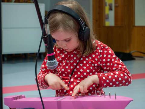 A five-year-old day-care centre pupil playing a pink traditional Finnish string instrument with headphones on. A microphone is recording the music.