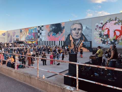 A band is performing for an audience in an industrial hall’s courtyard, the background shows graffiti art painted on the hall’s outer walls.