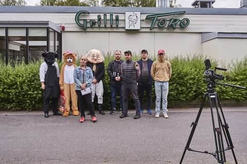 Ohjaaja, kuvaaja, kolme näyttelijää ja kolme kuvausryhmäläistä seisovat Grilli Toron kyltin alla. Videokamera etualalla.