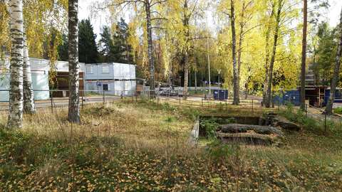 Autumnal birch trees have shed their leaves in the grassy courtyard with fortifications surrounded by fences. The old Jousenkaari School can be seen on the left. In the background is a sandy field with construction site containers visible.